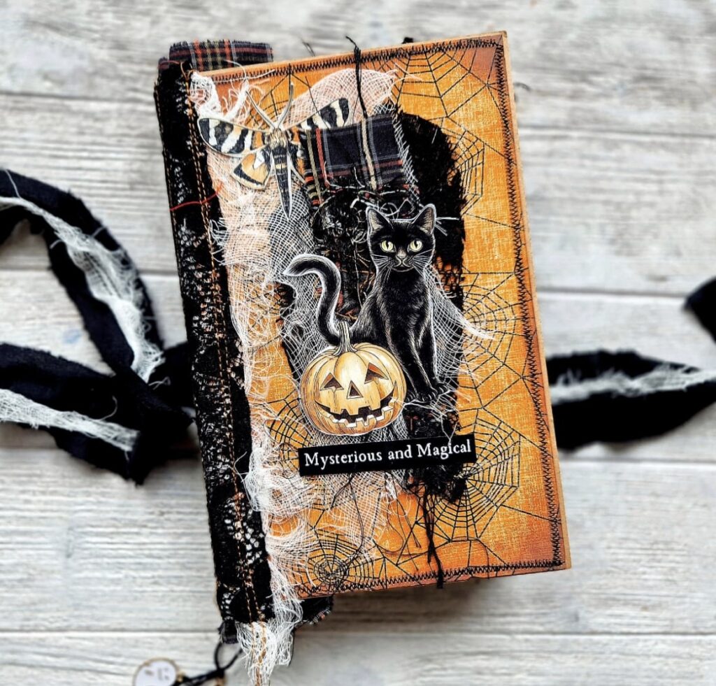 Halloween junk journal cover with a black cat, carved pumpkin, spiderweb background, and layered fabric textures