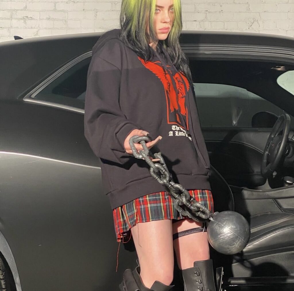 Billie Eilish with green hair, graphic hoodie, and ball and chain accessory. Creative inspiration for a Halloween Billie Eilish costume. Photo credit: @billieeilish on Instagram via Slowestuff.com