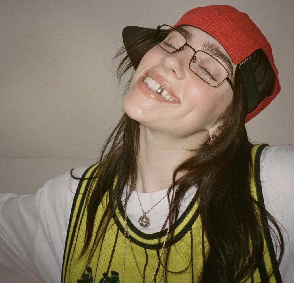 Billie Eilish wearing a red baseball hat and yellow jersey, showing her signature streetwear style and moody attitude. Photo credit: @billieeilish on Instagram via Slowestuff.com