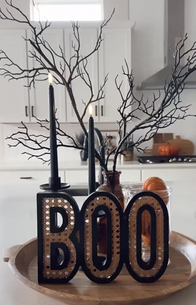 Rustic wood tiered tray styled with a “Boo” sign, twinkle lights, and orange beaded garland for a warm Halloween glow. Credit: mrs.alonzo.