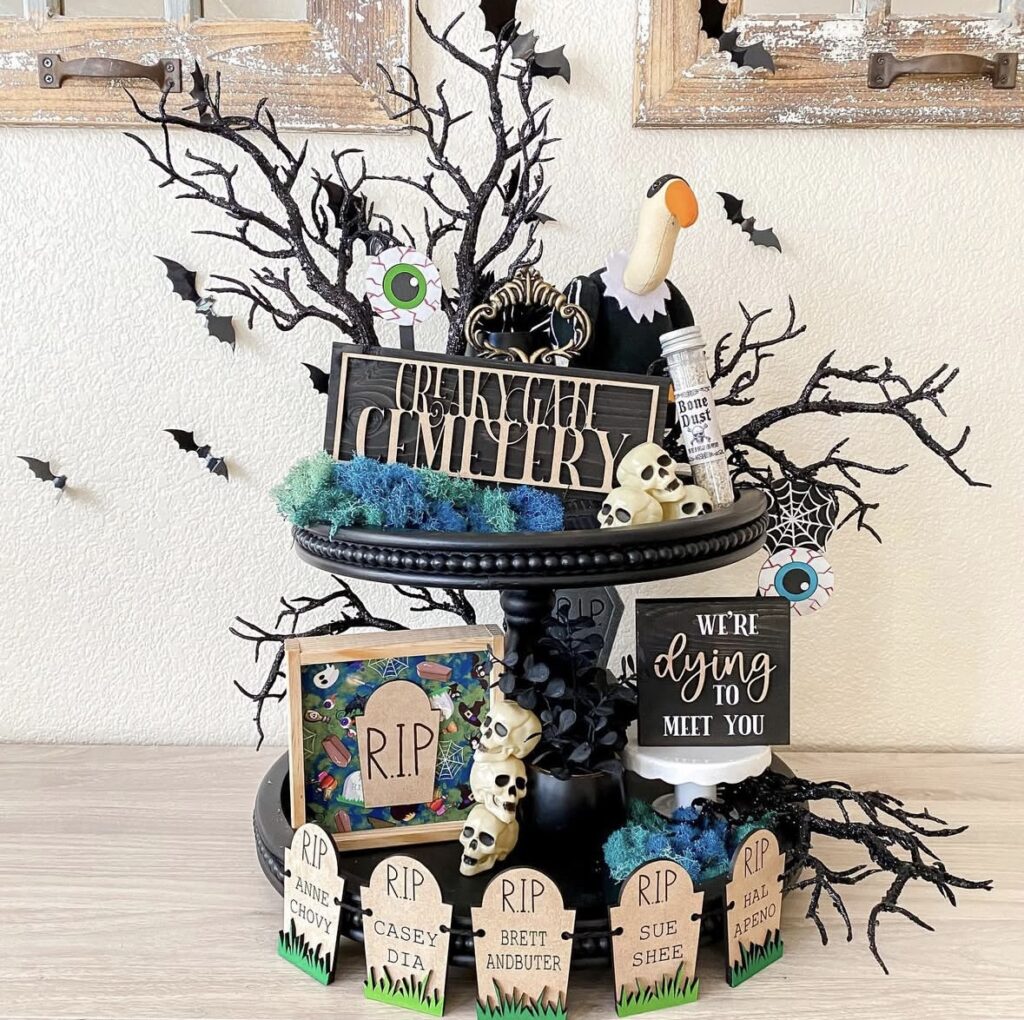 Skeleton-themed tiered tray decorated as a graveyard with tombstone signs, skulls, bone details, and spooky Halloween decor. Get skeleton tray ideas at www.slowestuff.com.