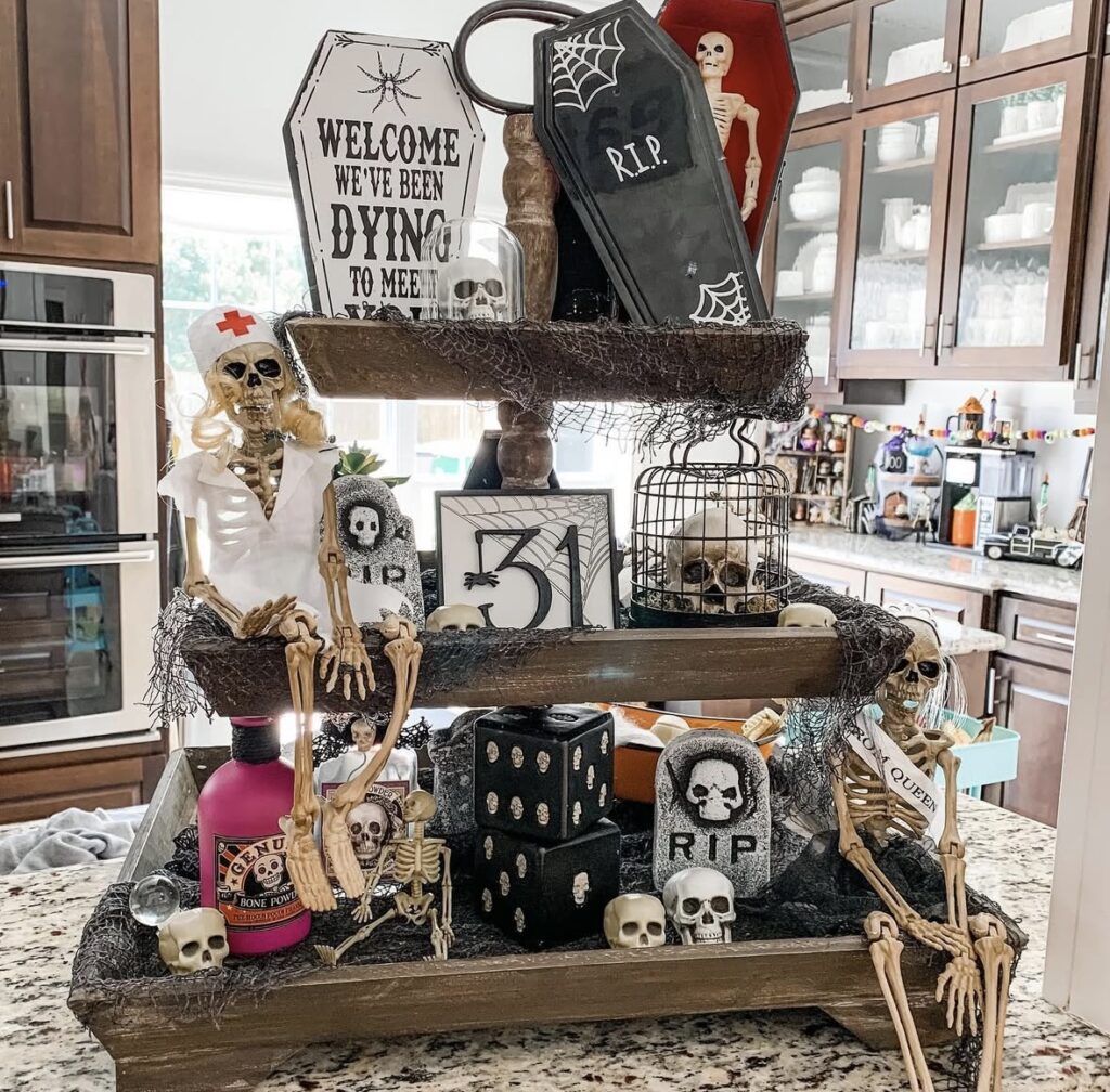 Tiered tray decorated for Halloween with mini skeletons, skull props, bone-shaped ornaments, and Halloween-themed signs. More skeleton tray ideas at www.slowestuff.com.