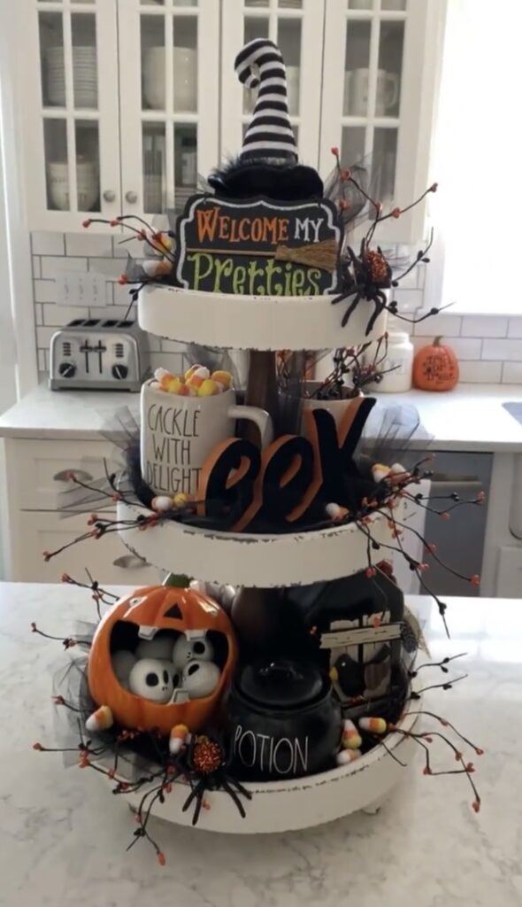 Halloween tiered tray with “Welcome My Pretties” sign, witch hat, black and white pumpkins, and orange accent decor. Credit: ondaisyhilldrive, www.slowestuff.com