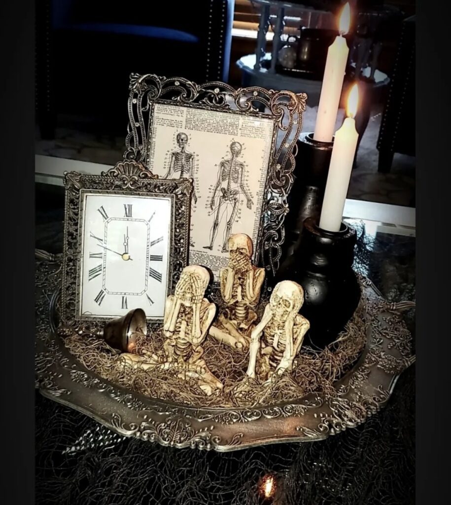 Halloween skeleton tray design with ornate metal tray, three skeletons doing see, hear and speak no evil, a framed clock and skeleton diagram with black candlestick holders and white skinny candles. Featured in slowestuff.com post on skeleton tiered tray designs