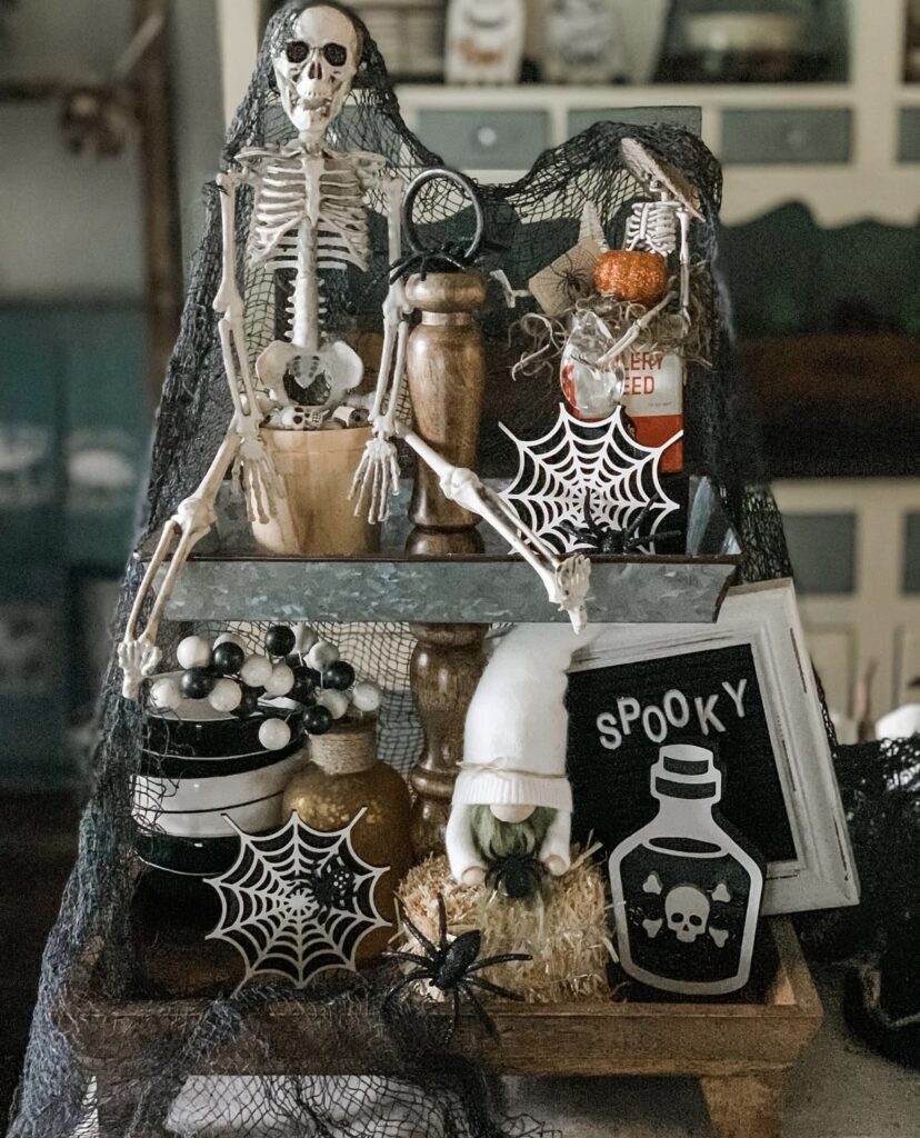 Skeleton tray decorated with creepy-cute details, including black netting over a skeleton, skull accents, spooky signs, and bone props for Halloween. Skeleton tiered tray ideas at www.slowestuff.com.
