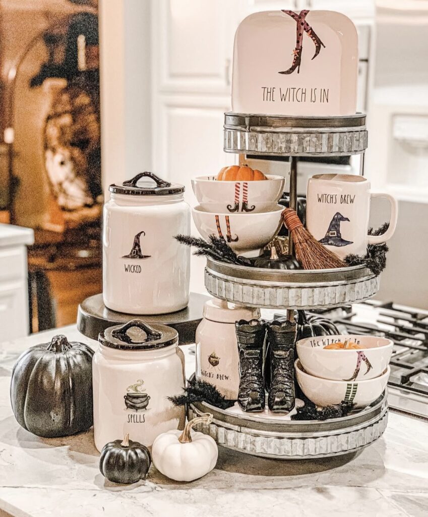 Black and white witch themed tiered tray with witch boots, tin tray, and a “The Witch Is In” sign for Halloween. Credit: jenna_design, www.slowestuff.com