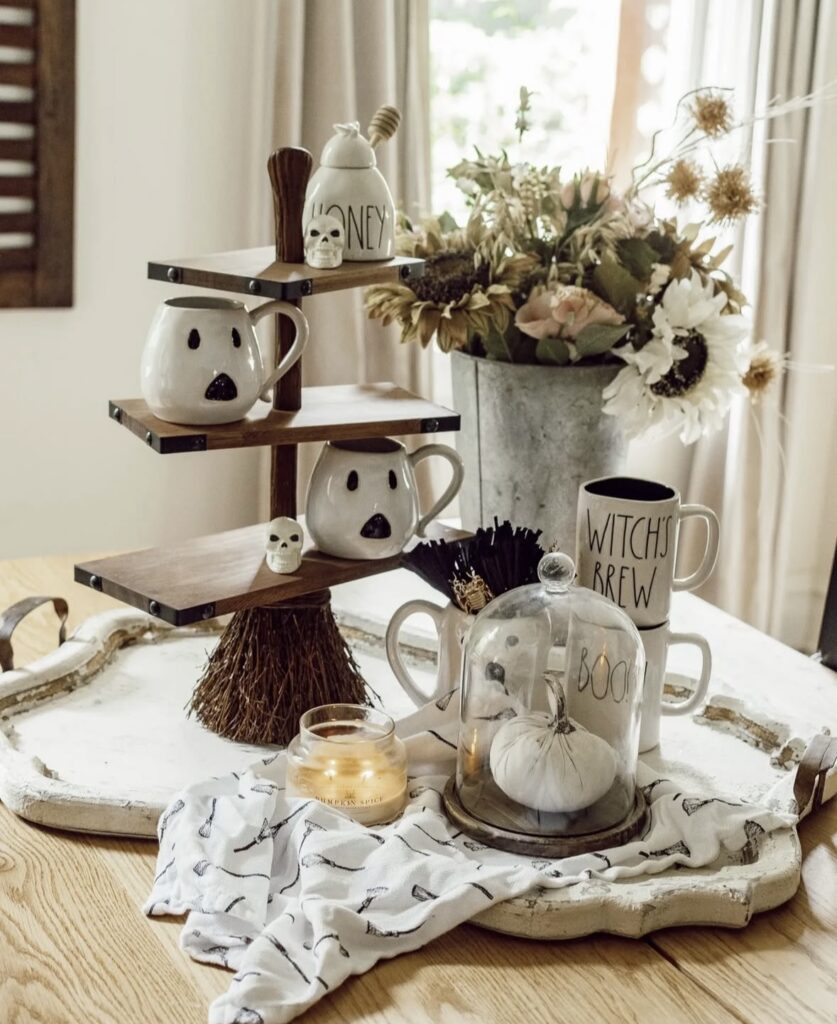 Wooden tiered tray styled like a witch’s broom, filled with cute ghost mugs and pops of black and white for a whimsical Halloween coffee bar. Credit: decorsteals and jamiewardtv.