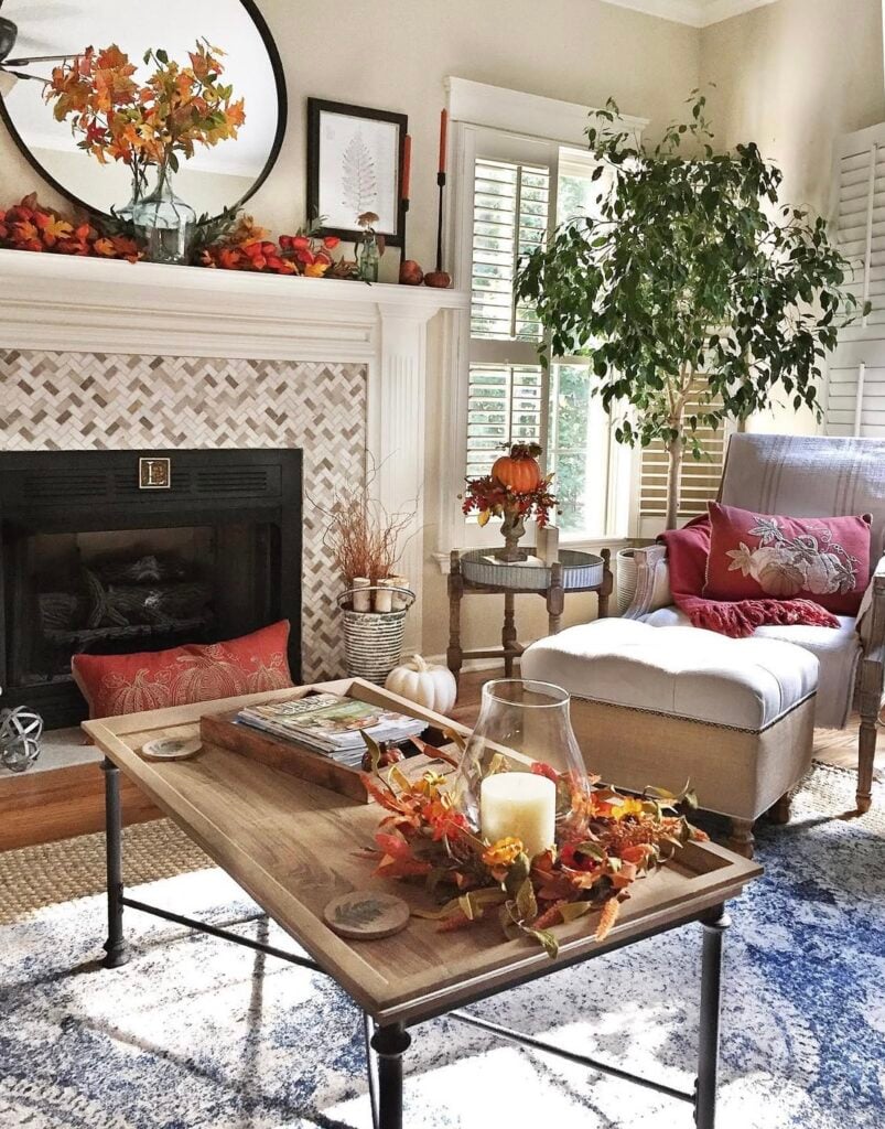 Cozy fall decor living room with leaf-garland mantel, round mirror, and a hurricane candle centerpiece on a wood coffee table