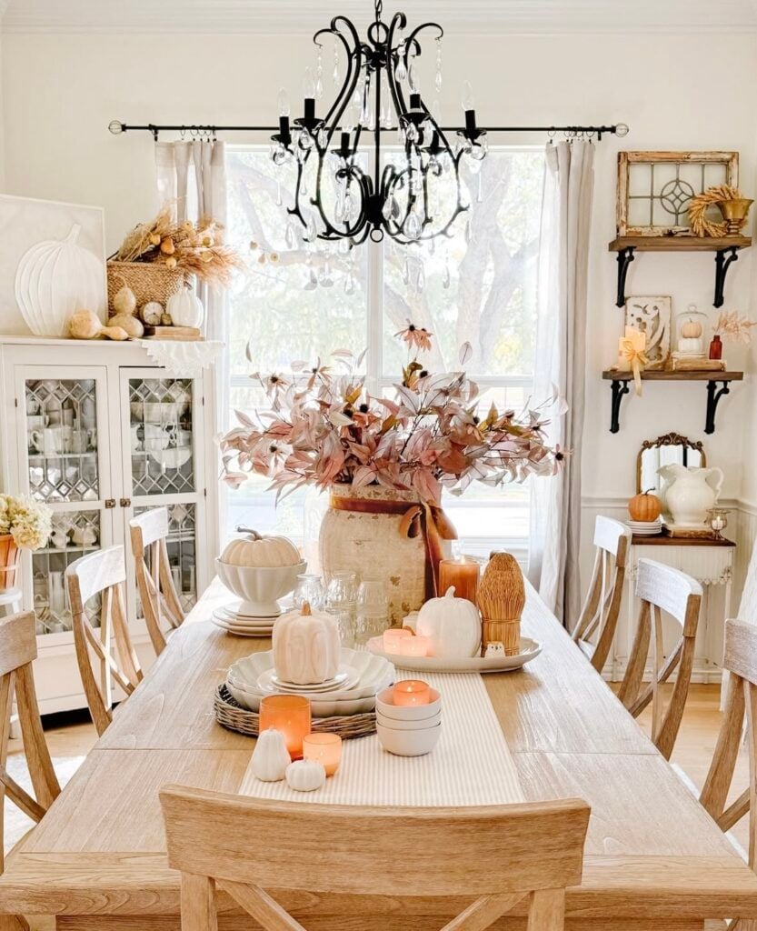 Fall dining table styled with blush foliage in a large vase, white ceramic pumpkins, stacked dishes, and glowing candles