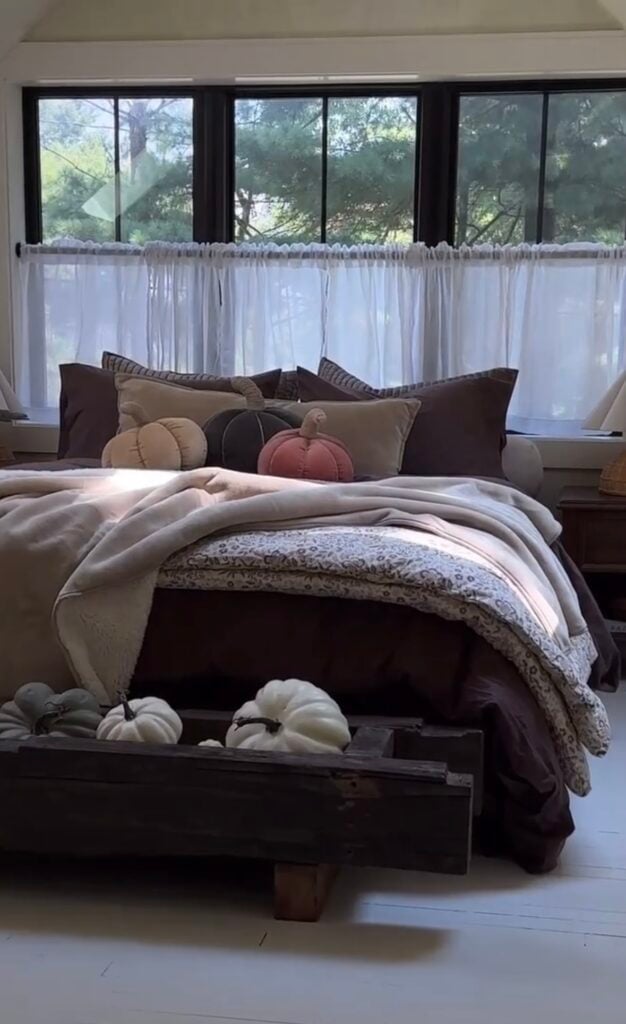 Brown and cream cozy fall bedroom with pumpkin pillows, layered bedding, and a rustic wooden tray filled with pumpkins at the foot of the bed.