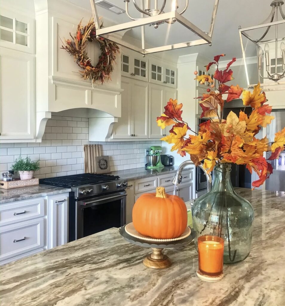 Colorful fall centerpiece on a kitchen island featuring orange pumpkins, faux florals, and autumn stems in a glass jar