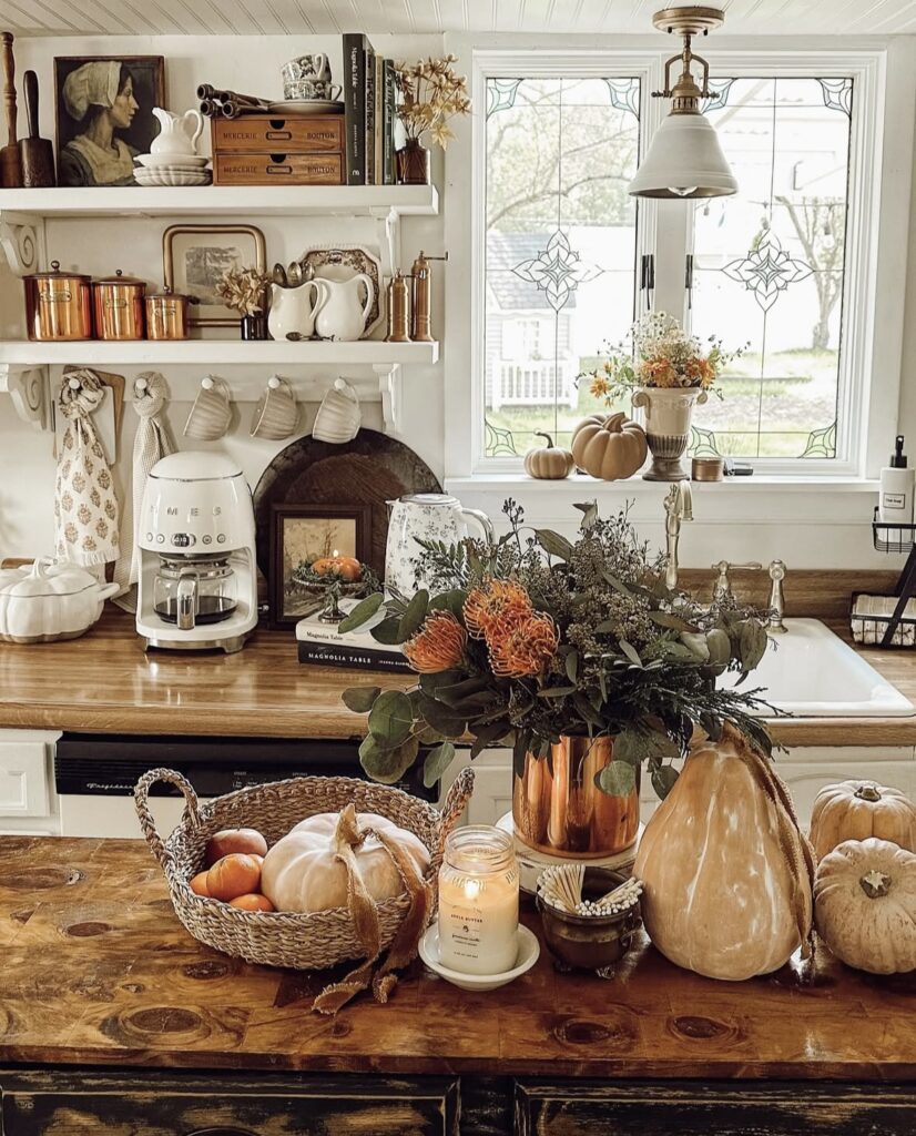 cozy white kitchen with shelves containing earthy accessories, wooden island with fall foliage and a bowl with pumpkins and other autumn accessories