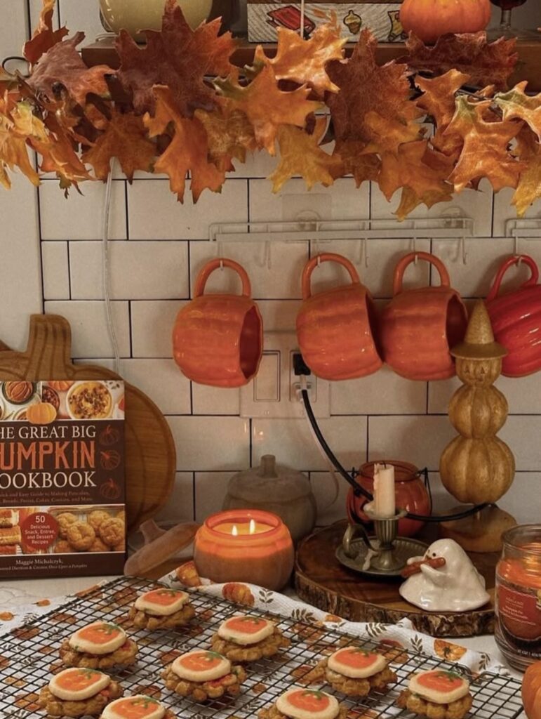Warm kitchen fall vignette with orange leaf garland, pumpkin cookbook, pumpkin mugs, and cooling rack of pumpkin cookies