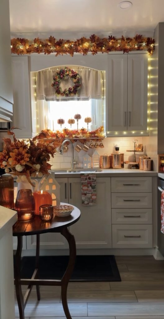 View of small white kitchen with fall garland around sink and small round table filled with fall foliage, twinkling lights and candles