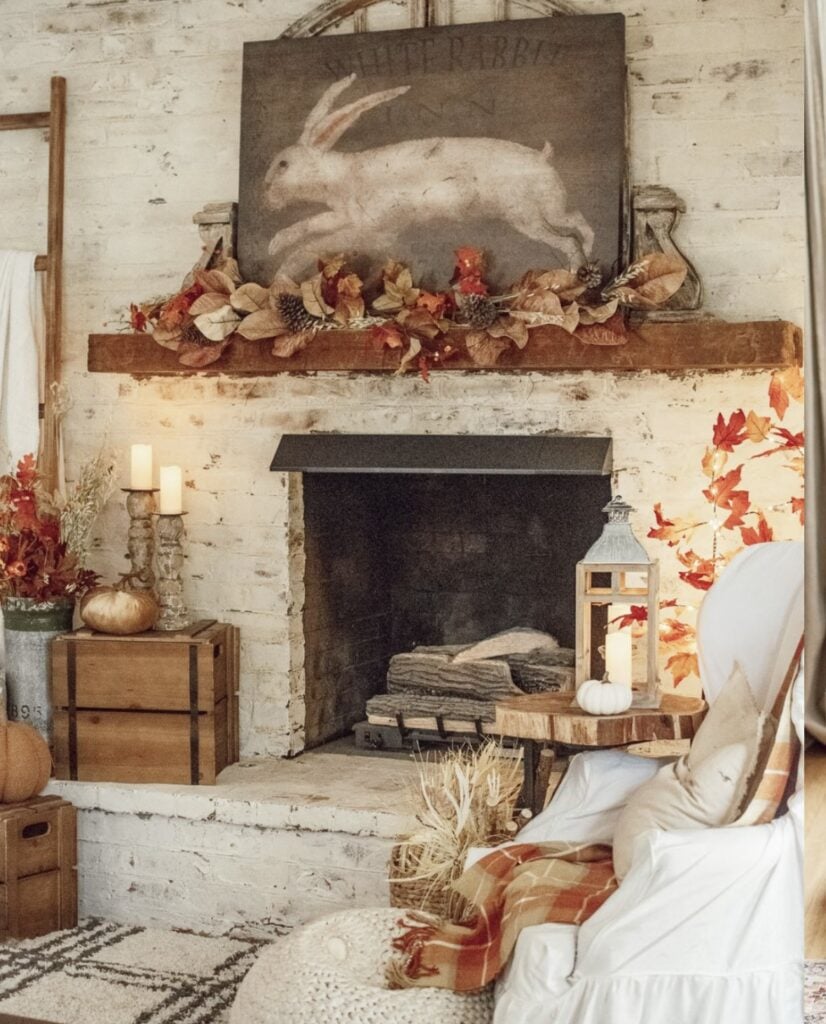 White brick fireplace with rabbit artwork, fall leaf garland, lantern and pumpkin; cozy chair with plaid throw