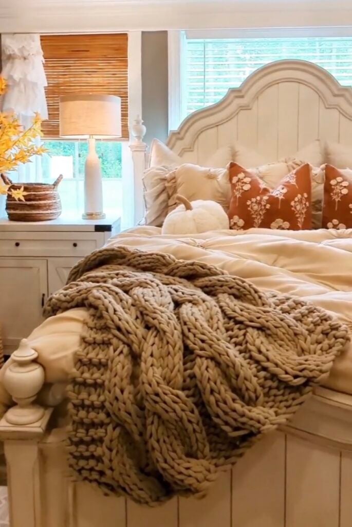 Cozy neutral fall bedroom with chunky knit blanket, burnt orange pillows, and white pumpkin accent for a warm autumn feel