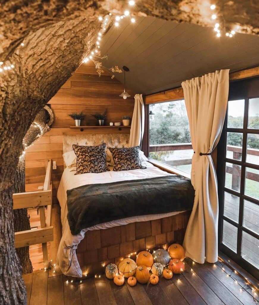 cozy treehouse bedroom with string lights, a bed dressed in white and dark green bedding, and a display of pumpkins on the floor