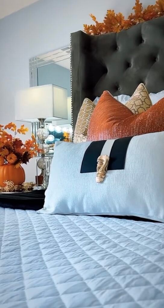 Pinterest pin graphic showing an elegant fall bedroom with a tufted headboard, orange throw pillows, autumn leaf decor, and a pumpkin floral arrangement