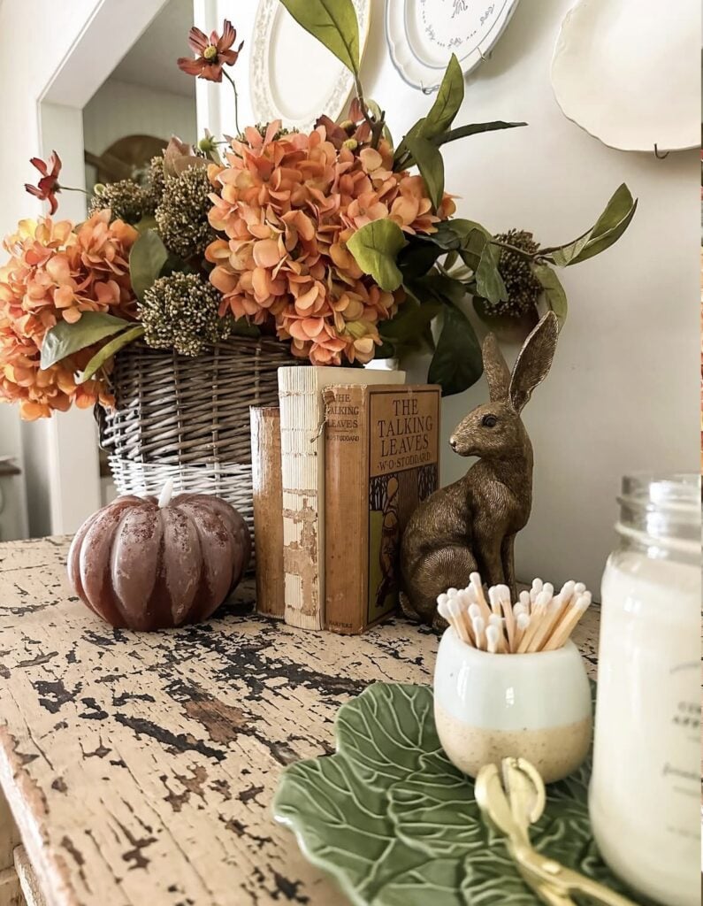 Entry table styled for fall with orange hydrangeas in a basket, vintage books, rabbit figurine, and a pumpkin candle
