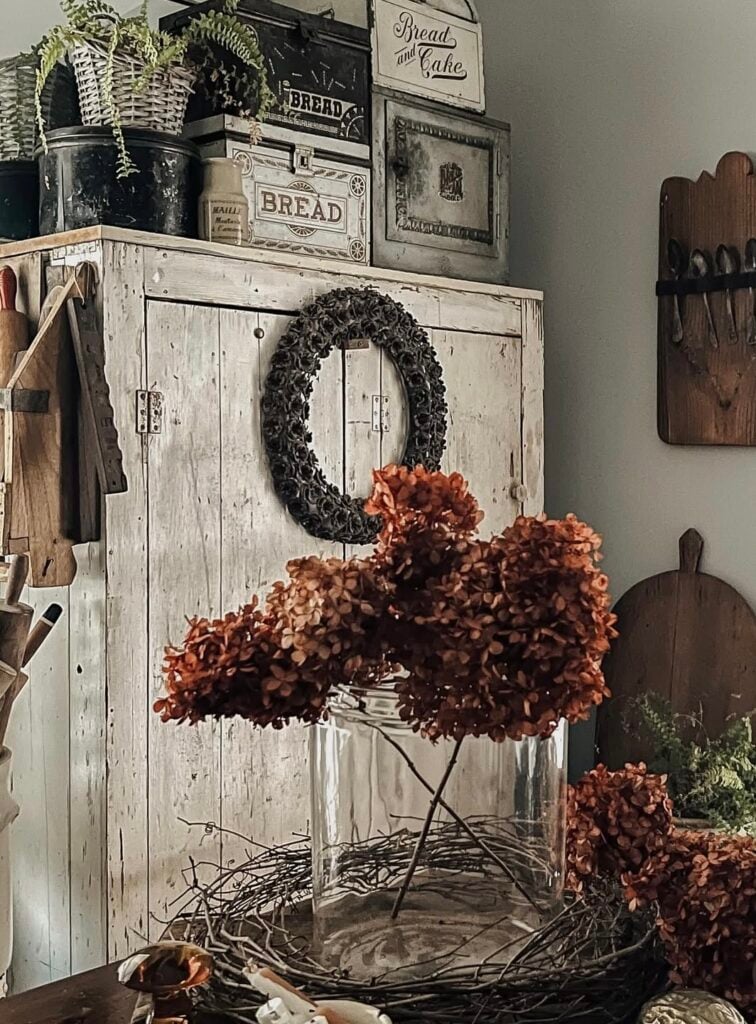 Moody farmhouse cabinet styled for fall with dried hydrangeas, wreath, and vintage bread tins.