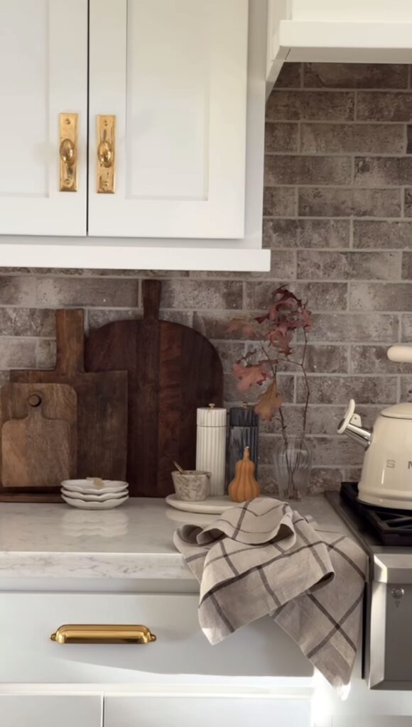 Kitchen counter with stone backsplash with dark wood cutting board propped up, candle and a small pear in fall orange color
