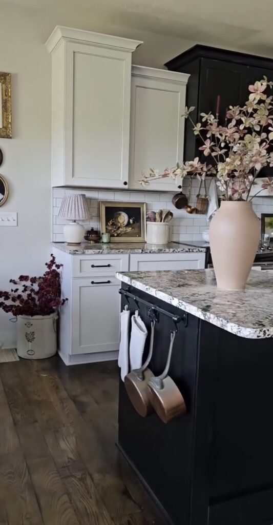 White kitchen with dark island with vase and autumn foliage with vignette of artwork, jars, lamp and candles