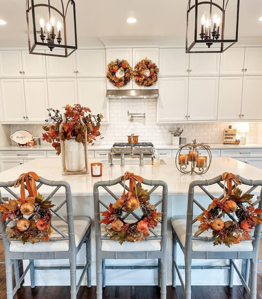 Bright white kitchen decorated for fall with wreaths on cabinets, orange floral arrangement on island, and mini pumpkin wreaths on bar stools