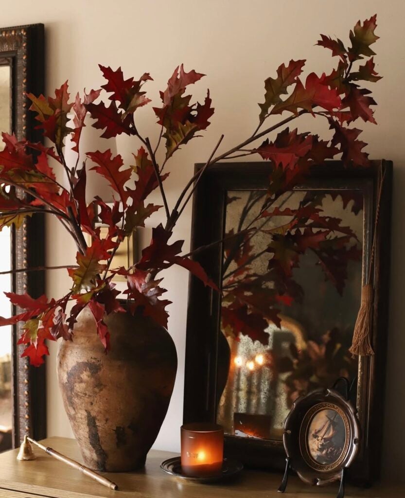Large clay vase of red oak branches beside an antique mirror with a lit amber candle—warm fall living room vignette.