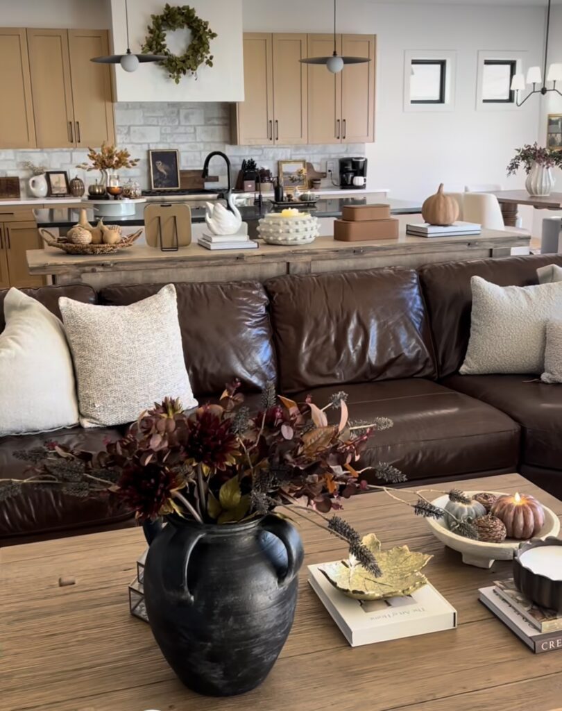 Open-concept living room with brown leather sofa and coffee table styled with moody fall florals and pumpkins.