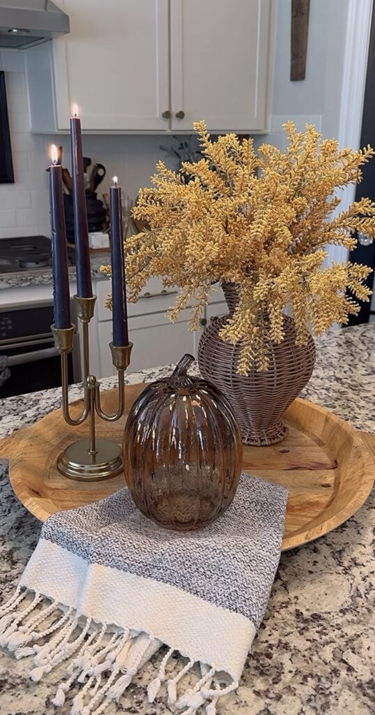 Wooden tray with dark taper candles, wicker vase of golden foliage, glass pumpkin, and fringed towel on granite countertop.