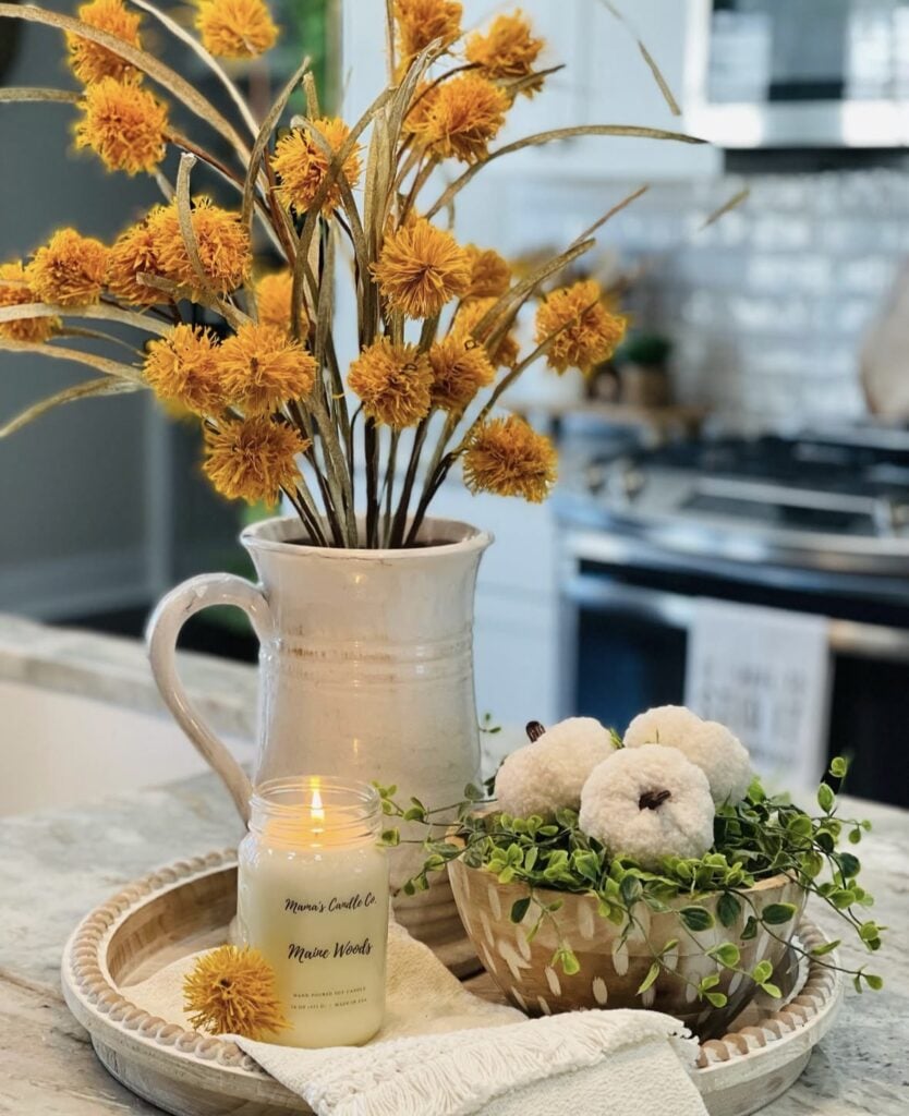 Light wooden tray with cream pitcher of yellow pom-pom flowers, lit candle, and wooden bowl of white pumpkins with greenery