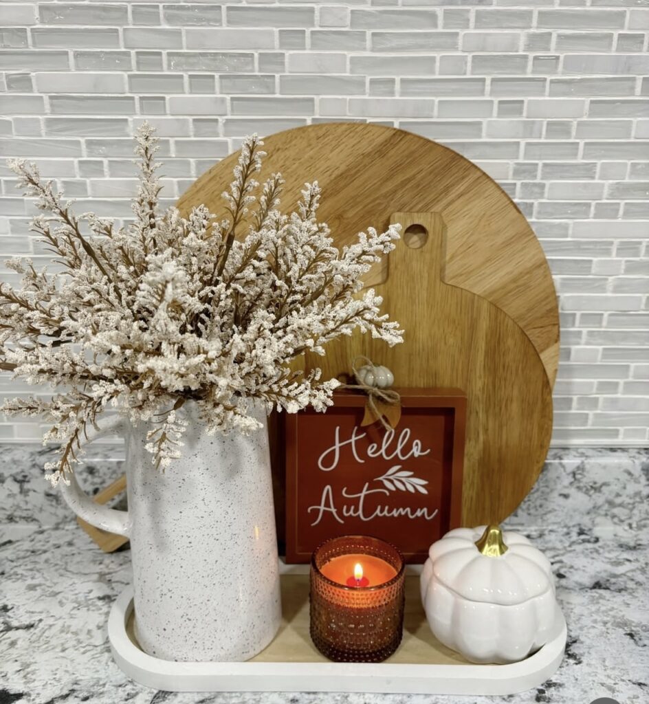 Small white tray with speckled pitcher of frosted foliage, lit candle, white ceramic pumpkin, and wooden cutting boards