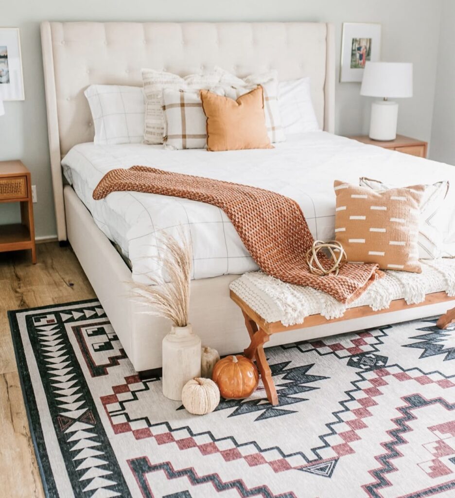 Modern fall bedroom with a beige upholstered bed, white and tan plaid bedding, burnt orange throw blanket, and decorative pumpkins on a patterned rug.