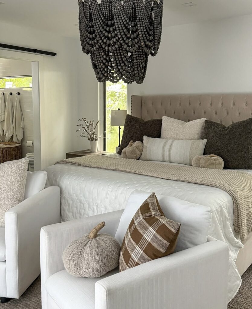 Neutral fall bedroom with a tufted headboard, layered beige bedding, plaid throw pillow, and knit pumpkin decor on white chairs.