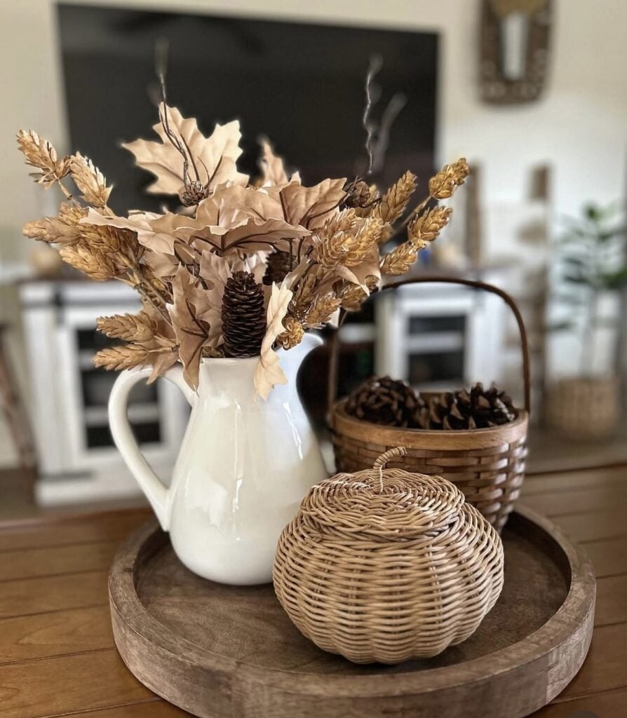 Round wooden tray with white pitcher of neutral foliage, wicker pumpkin, and basket of pinecones