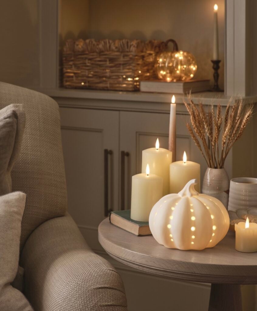Cozy neutral side table with glowing pillar candles, ceramic pumpkin luminary, stacked books, wheat stems in a bud vase, and woven basket with twinkle lights