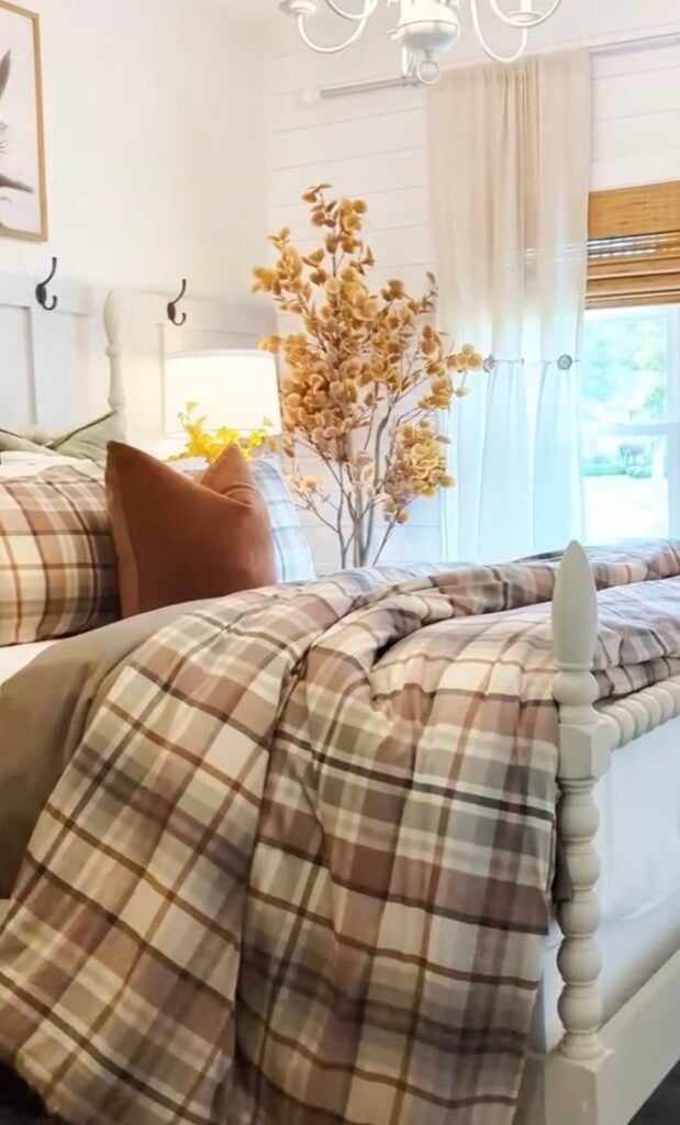 Farmhouse-style fall bedroom with plaid bedding, rust-colored throw pillows, and a tall vase of autumn foliage.