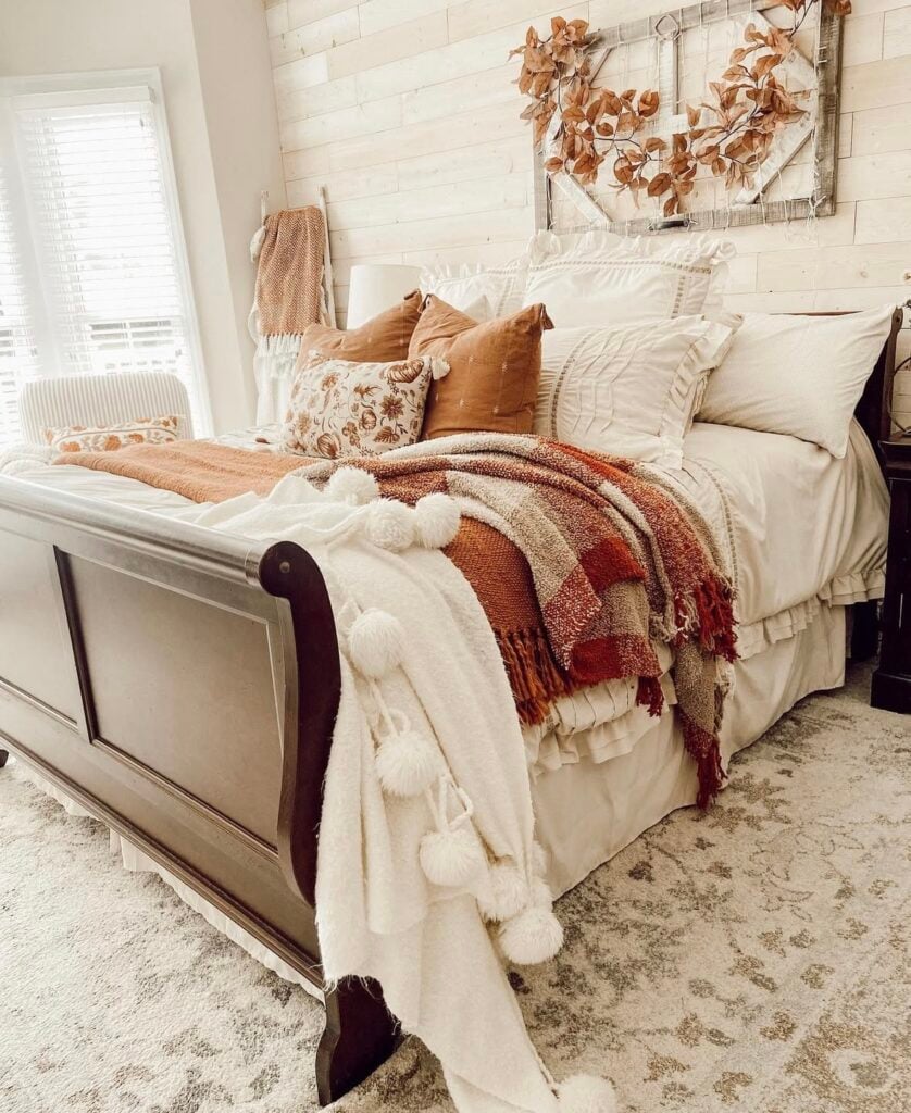 Warm farmhouse-style fall bedroom with a dark wood sleigh bed, cream bedding, orange pillows, and a plaid throw blanket, accented by a leaf wreath above the headboard.