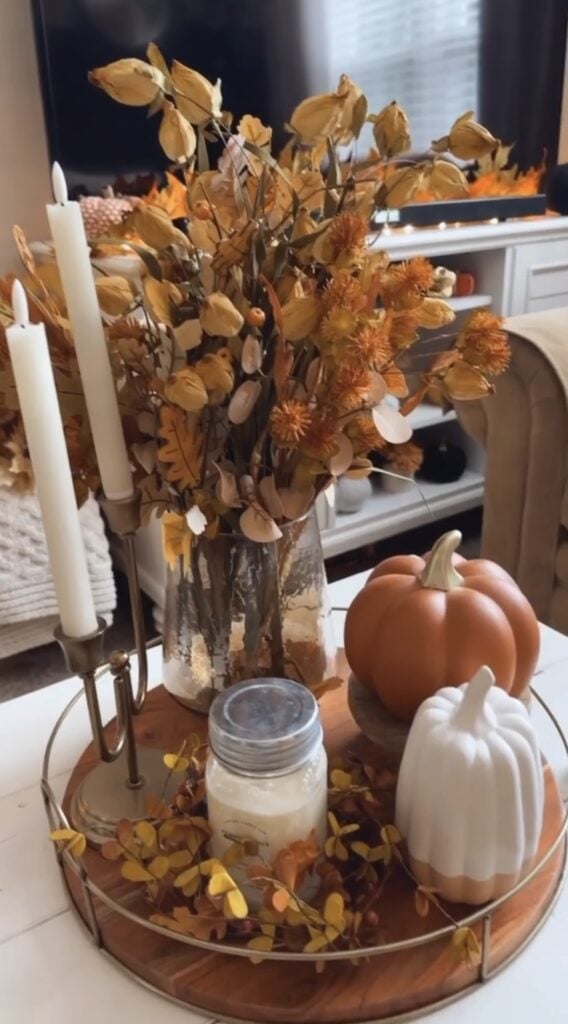 Round tray with tall candles, amber vase of yellow flowers, ceramic pumpkins, and a jar candle.