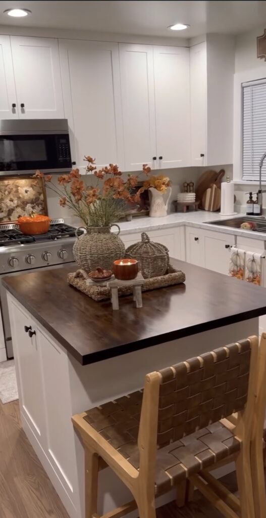 White kitchen with small island with dark countertop, woven tray, vase and pumpkin with fall stems and orange color accents