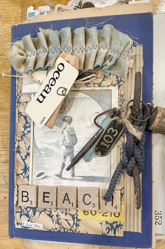 Vintage beach junk journal cover with a blue background, layered ephemera, a vintage boy-at-the-shore photo, keys and a “103” tag, an “ocean” label, and Scrabble tiles spelling “BEACH.”