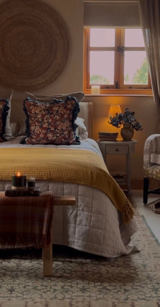 Inviting autumn bedroom with soft lighting, neutral quilt, mustard blanket, and decorative floral pillows, styled with warm wood accents and a large woven wall hanging.