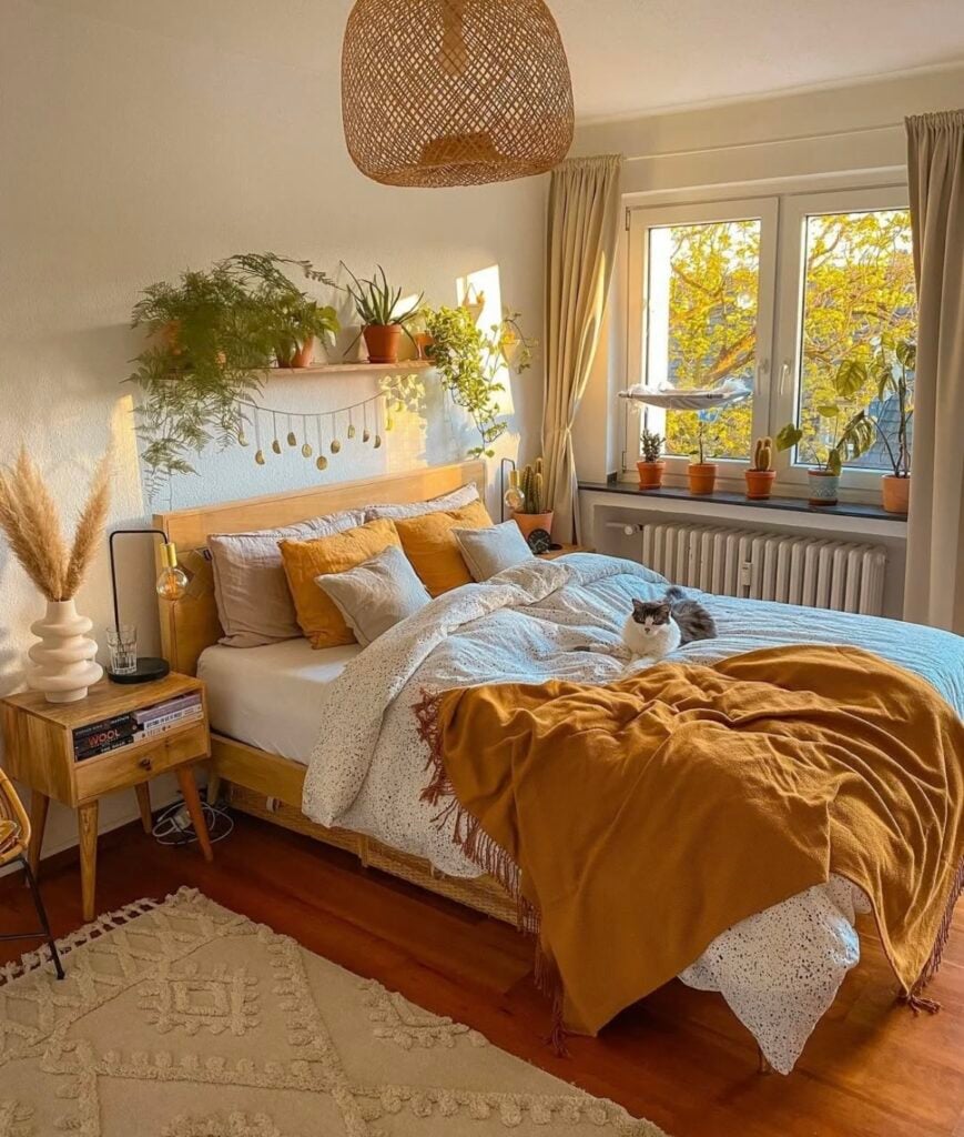 Bright and cozy fall bedroom with mustard throw blanket, neutral bedding, and a mix of tan and gray pillows, with potted plants along the window and a cat lounging on the bed.
