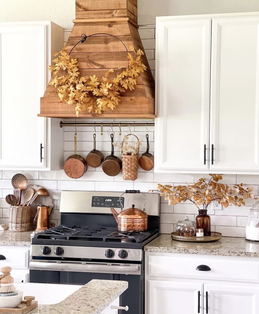 White kitchen with wooden vent over stove with copper cookware with amber bottles filled with fall foliage