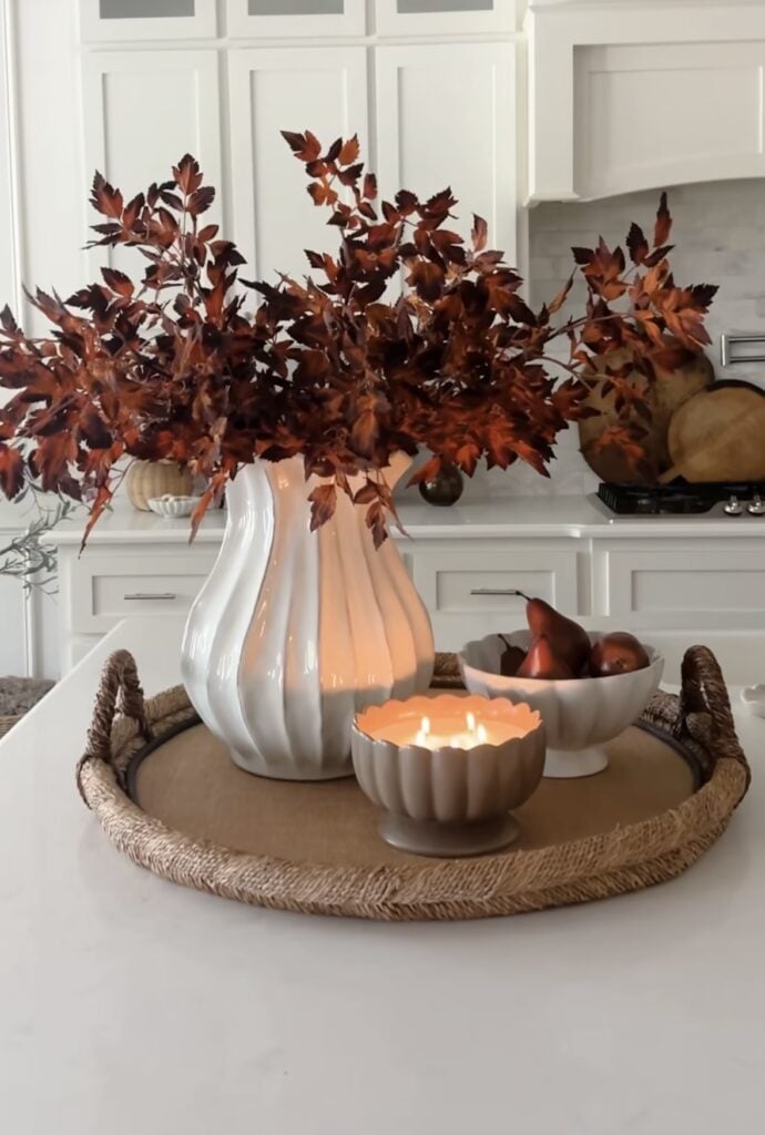 Woven tray with white fluted vase of deep red leaves, lit candle, and small bowl of pears