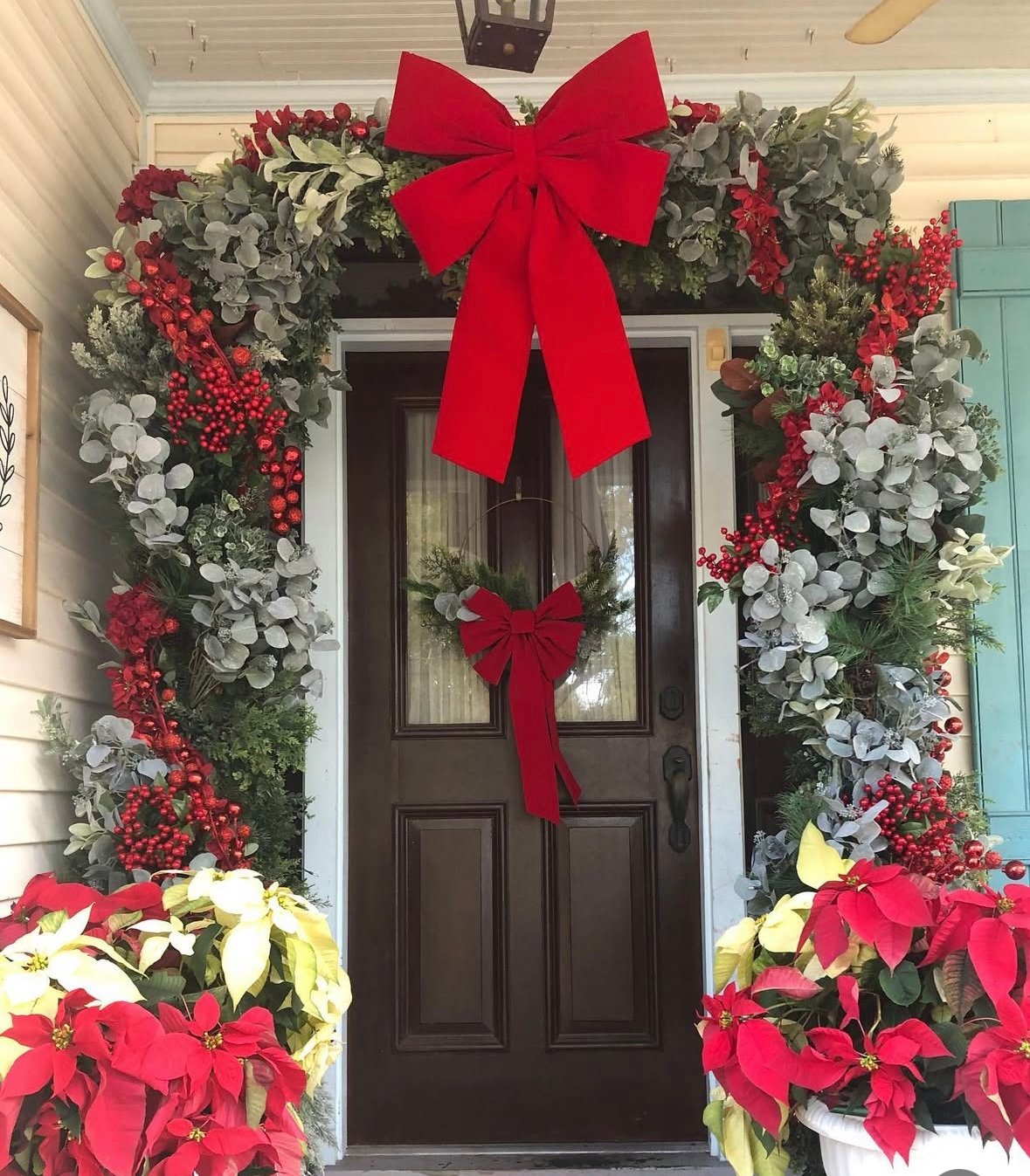 Classic Christmas front door framed in lush evergreen and eucalyptus garlands with bright red berries, huge red bow at the top, matching ribboned wreath, and planters of red and cream poinsettias on a black door