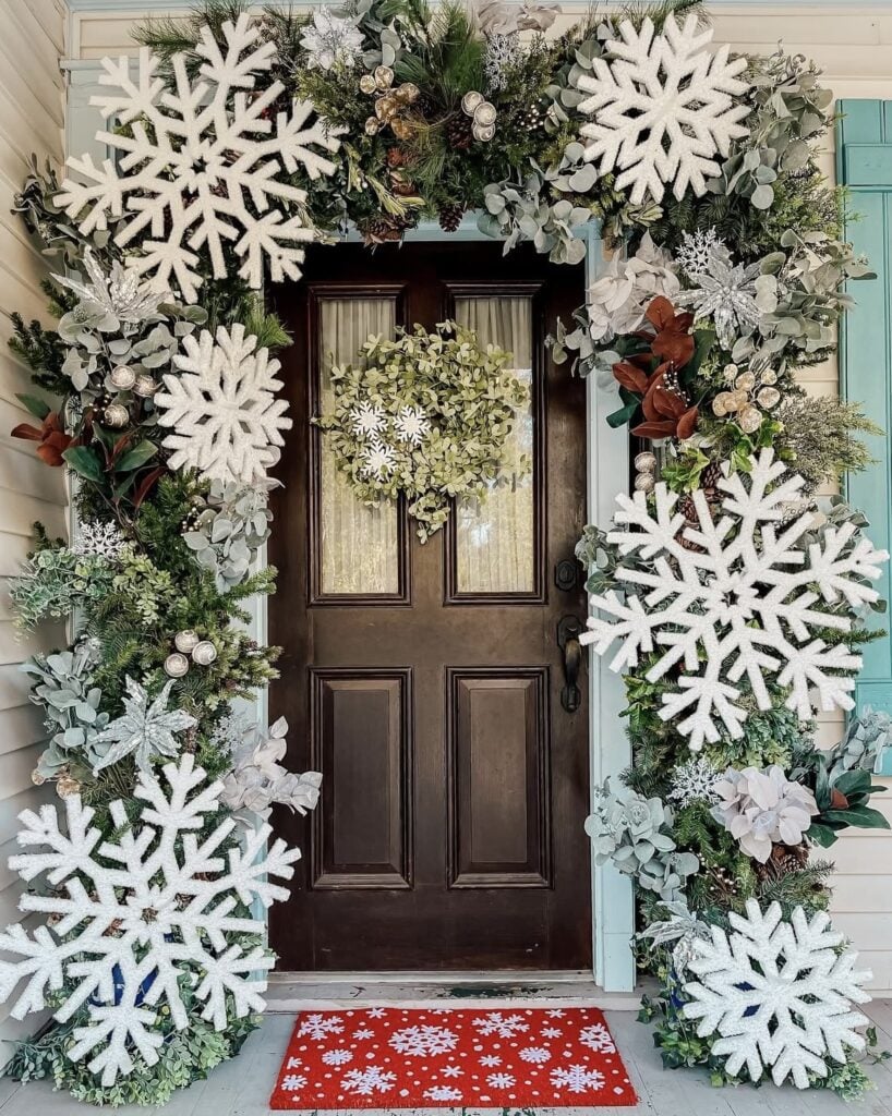 Serene woodland Christmas entry with layered greenery, pine cones, magnolia leaves, and oversized white snowflakes around a simple snowflake wreath
