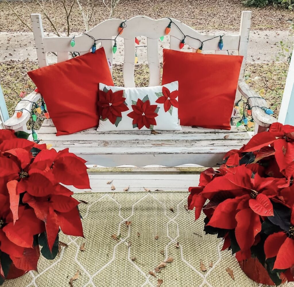 Cozy porch bench styled for Christmas with red and white pillows, poinsettia accents, and warm string lights for a classic holiday look