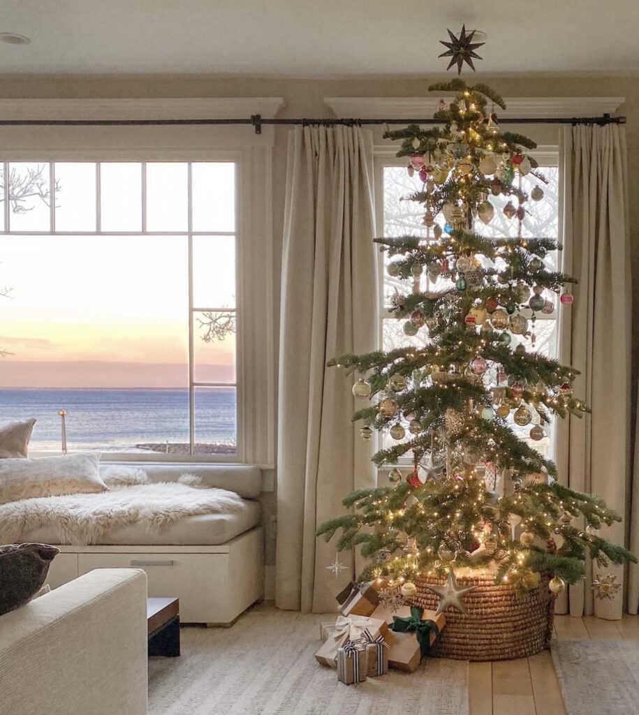 coastal living room with slim tree in basket collar, vintage glass ornaments, warm white lights, large picture window at sunset