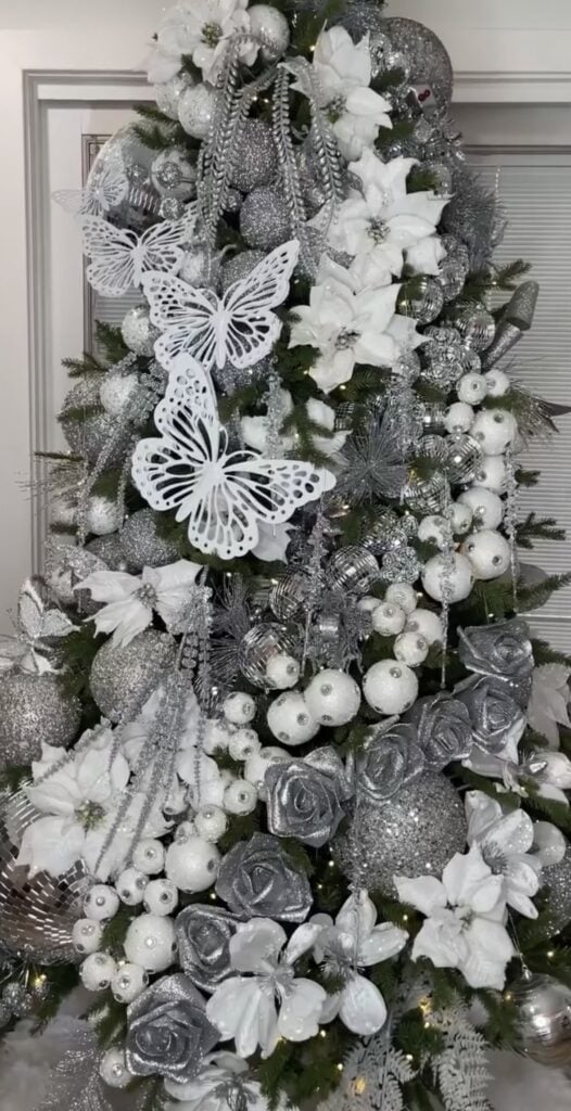 High-glam white and silver Christmas tree—giant cutout butterflies, white poinsettias and roses, disco-ball ornaments, pearl garlands, and icy sprays for a luxe, overflowing look.